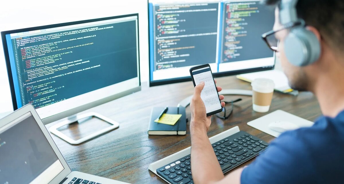 Man Viewing Code On Mobile With Computer Behind