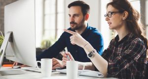 Woman Pointing At Monitor While Taking Notes, Man Typing